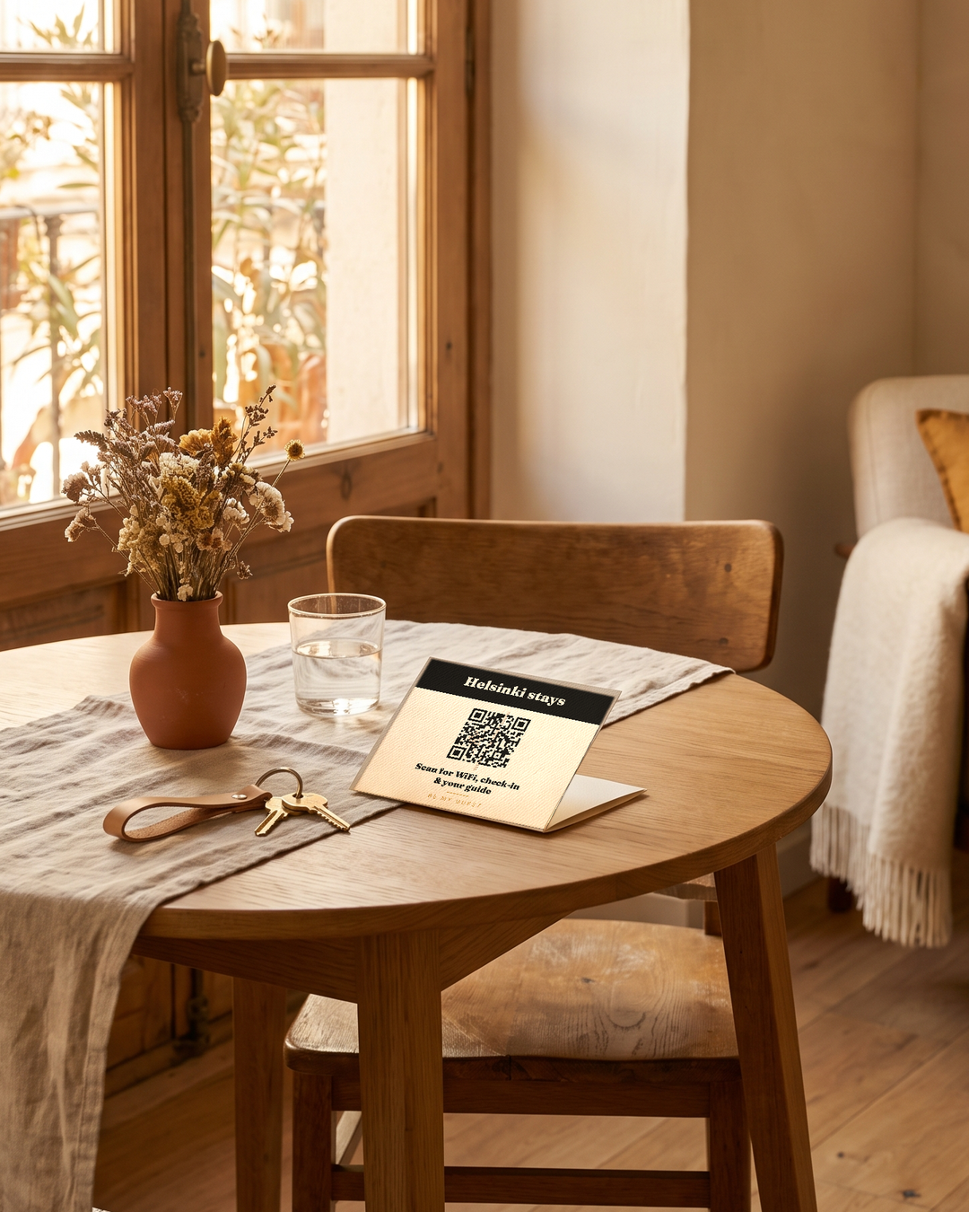 A printed QR card standing on an apartment table beside keys, a glass of water and dried flowers, reading "Scan for WiFi, check-in & your guide".